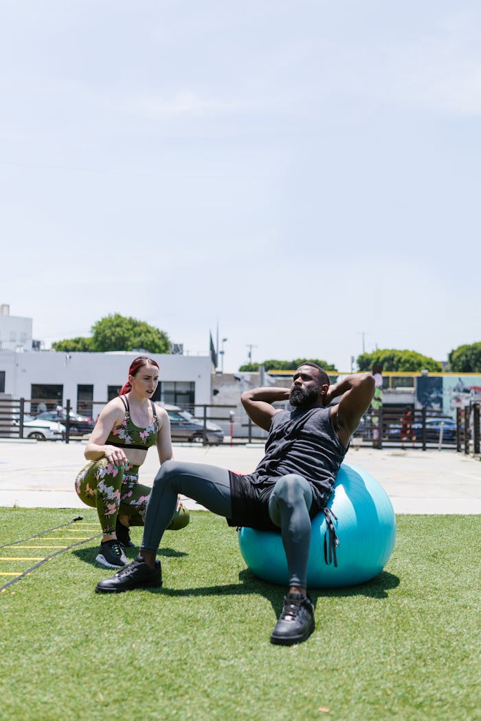 A man doing sit-ups on an exercise ball outdoors with a trainer guiding him, promoting fitness.